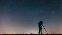 A silhouetted person using a telescope to observe the night sky filled with stars.