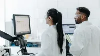 Two scientists wearing lab coats and goggles analyze data on a computer in a modern laboratory.