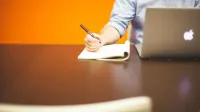 Adultos escribiendo notas en un cuaderno junto a una computadora portátil en un escritorio de madera con fondo naranja.