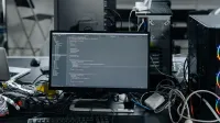 A cluttered workstation in an office featuring a monitor displaying code, surrounded by a keyboard, mouse, and wiring.