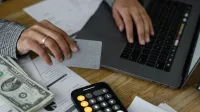 Close-up of a person using a credit card and laptop in a financial setting with cash and calculator.