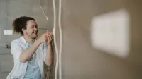An electrician with curly hair adjusts an outlet indoors, smiling as he works.