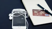 A white smartphone with earbuds, credit card, and pen on a notepad, symbolizing tech and finance.