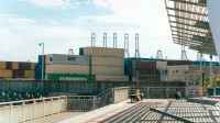 Vista de un puerto de envío con contenedores apilados y un hombre descansando cerca bajo sombras industriales.