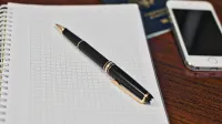 Black pen on a grid notebook with smartphone and passport on wooden desk.