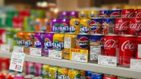 Coloridas latas de refresco que incluyen Fanta, Pepsi y Coca-Cola dispuestas en los estantes de los supermercados, etiquetas de precio visibles.