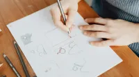 Desde arriba de una diseñadora femenina sin rostro en suéter de punto sentado en una mesa de madera con una hoja de papel mientras dibuja con lápiz