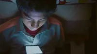 Boy lying in bed staring intently at a glowing smartphone screen.