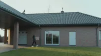 Person delivering package to a modern brick house with a tile roof and green lawn.
