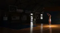 Un hombre practica tiros de baloncesto en una cancha cubierta con poca luz, creando una atmósfera dramática y enfocada.