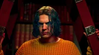 Kai Anderson (Evan Peters) mira hacia adelante con una expresión intensa, enmarcado por una estructura roja con una placa de metal que muestra el número 13; usa un suéter naranja de punto