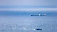 Un barco de carga y un submarino navegan por el vasto océano abierto bajo un cielo despejado.