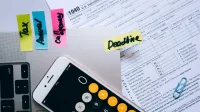 Office desk flat lay showing tax documents, calculator app on smartphone, sticky notes, and paperclips.