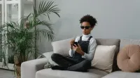 Un adolescente con discapacidad visual sentado en un sofá usando un smartphone y auriculares.