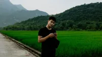 Hombre asiático de pie con smartphone en un paisaje rural brumoso, rodeado de campos de arroz verde y montañas.