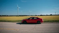 Un sedán rojo en un camino rural junto a campos con turbinas de viento bajo un cielo azul claro.