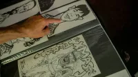 Primer plano de una mano apuntando a los bocetos de caricatura en una cartera de arte sobre una superficie oscura.