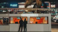 Dos personas de pie en una taquilla bajo luces de neón por la noche, entorno de cine urbano.