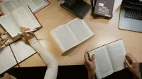 Grupo de adultos estudiando junto con libros abiertos y portátiles en una mesa de madera.