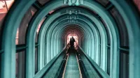 Un túnel futurista con una escalera mecánica iluminada y siluetas de personas, mostrando la arquitectura moderna.