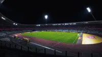Estadio de fútbol vacío brillantemente iluminado con campo verde bajo los focos durante la noche.