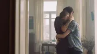 A loving embrace between two men in a softly lit room, showcasing warmth and affection.