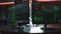 Close-up of dual computer monitors with green coding interfaces in a dark room, highlighting cyber security themes.