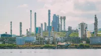 Un gran complejo de refinería industrial con chimeneas altas junto al mar bajo un cielo despejado.