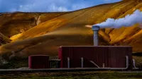 Planta de energía geotérmica roja que emite vapor contra un dramático telón de fondo montañoso de color amarillo-marrón.