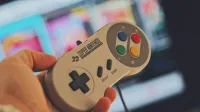 Hand holding a classic Super Nintendo game controller indoors with a blurred screen in the background.