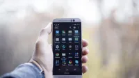 A close-up of a hand holding an HTC smartphone with visible apps on the screen, in an outdoor setting.