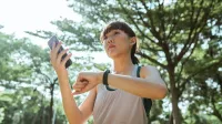 Desde abajo étnica mujer asiática con ropa deportiva de pie en el parque y conectando smartphone y reloj inteligente durante el descanso en día soleado