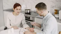 Adult man monitoring elderly woman's blood pressure at kitchen table using sphygmomanometer.