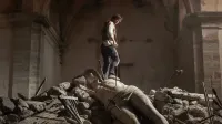 Una persona se encuentra sobre escombros dentro de un viejo edificio de piedra dañado, con una gran estatua rota que se encuentra entre los escombros, creando una atmósfera dramática y