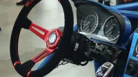 Close-up view of a custom racing steering wheel with red accents, featuring a dashboard and speedometer in a racing setting.