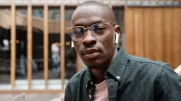 Hombre afroamericano calvo con gafas y auriculares, con una mirada segura, al aire libre.