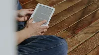 Close-up de manos sosteniendo un e-reader en un piso de madera, representando un momento de lectura casual.