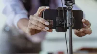 Crop unrecognizable female setting mobile phone on tripod while recording vlog on blurred background