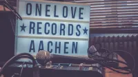 A warm-toned studio featuring a microphone, audio mixer, and lightbox signage reading 'One Love Records Aachen'.