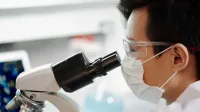Científico con máscara facial y gafas examinando muestras a través de un microscopio en un laboratorio.