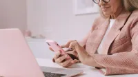Anciana en traje de cuadros rosa con un smartphone y una computadora portátil en un escritorio.