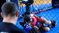 Two women competing in a mixed martial arts training session inside a gym cage with a coach observing.