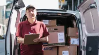 El repartidor de edad avanzada tiene portapapeles frente a una furgoneta abierta llena de paquetes en un día soleado.
