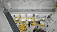 Área común de prisión con múltiples reclusos con uniformes amarillos tirados en el suelo, vistos desde arriba. Paredes, puertas, mesas y escaleras blancas en un entorno seguro. cámara de