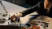 Un detective ajustando una grabadora durante una investigación con fotos esparcidas sobre una mesa.