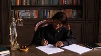 Un juez vestido de túnica escribiendo en un documento en un escritorio en una biblioteca de oficina con libros de derecho.