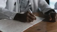 Primer plano de un médico escribiendo notas con un bolígrafo en un cuaderno médico en un escritorio de madera.