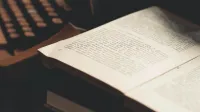 An open book with visible text in front of an antique typewriter, evoking a vintage literary theme.