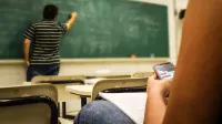 Student texting in a classroom while teacher is writing on the blackboard.