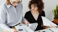 Two women review and discuss financial documents in an office setting, highlighting teamwork and analysis.